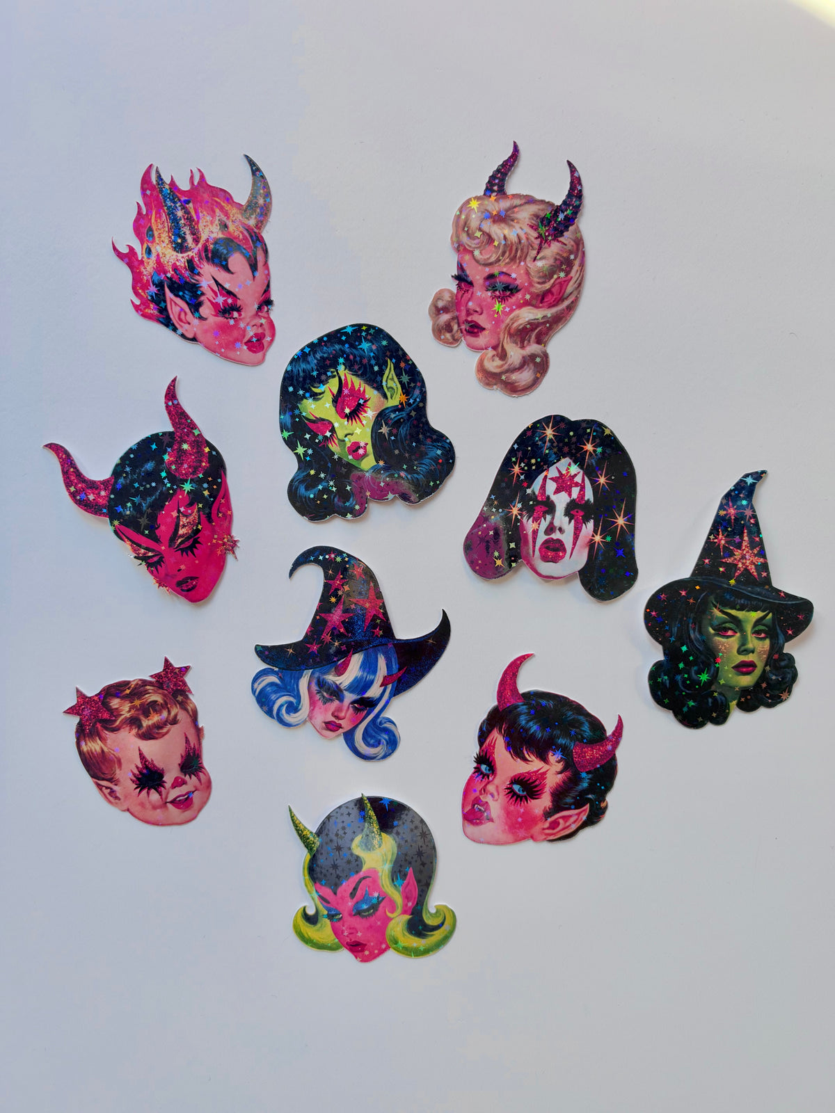 A set of nine colorful witch and devil-themed hair clips arranged on a plain light gray background. The clips feature various designs including stars, sparkles, and cosmic patterns in pink, blue, green, and black.
