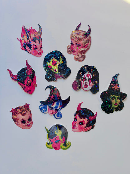 A set of nine colorful witch and devil-themed hair clips arranged on a plain light gray background. The clips feature various designs including stars, sparkles, and cosmic patterns in pink, blue, green, and black.