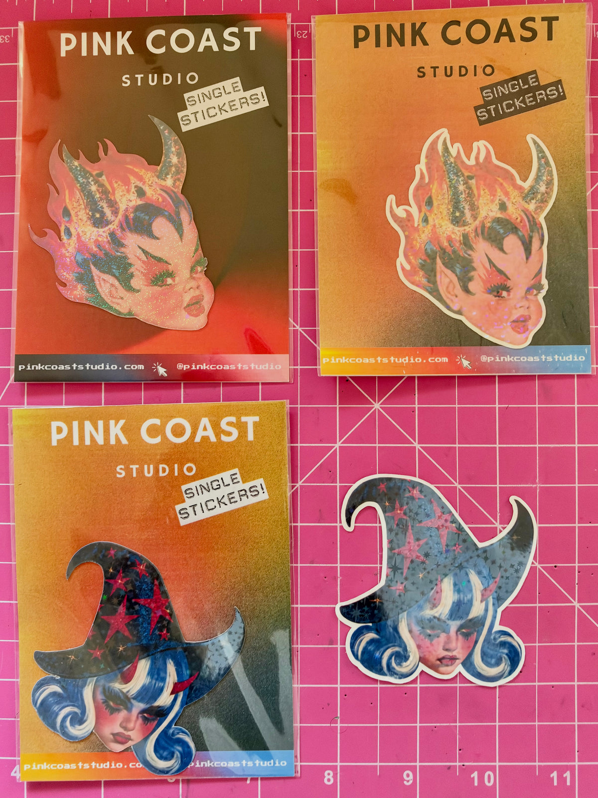 Four Pink Coast Studio single stickers displayed on a pink cutting mat background, featuring stylized horned and witch-themed illustrated faces.