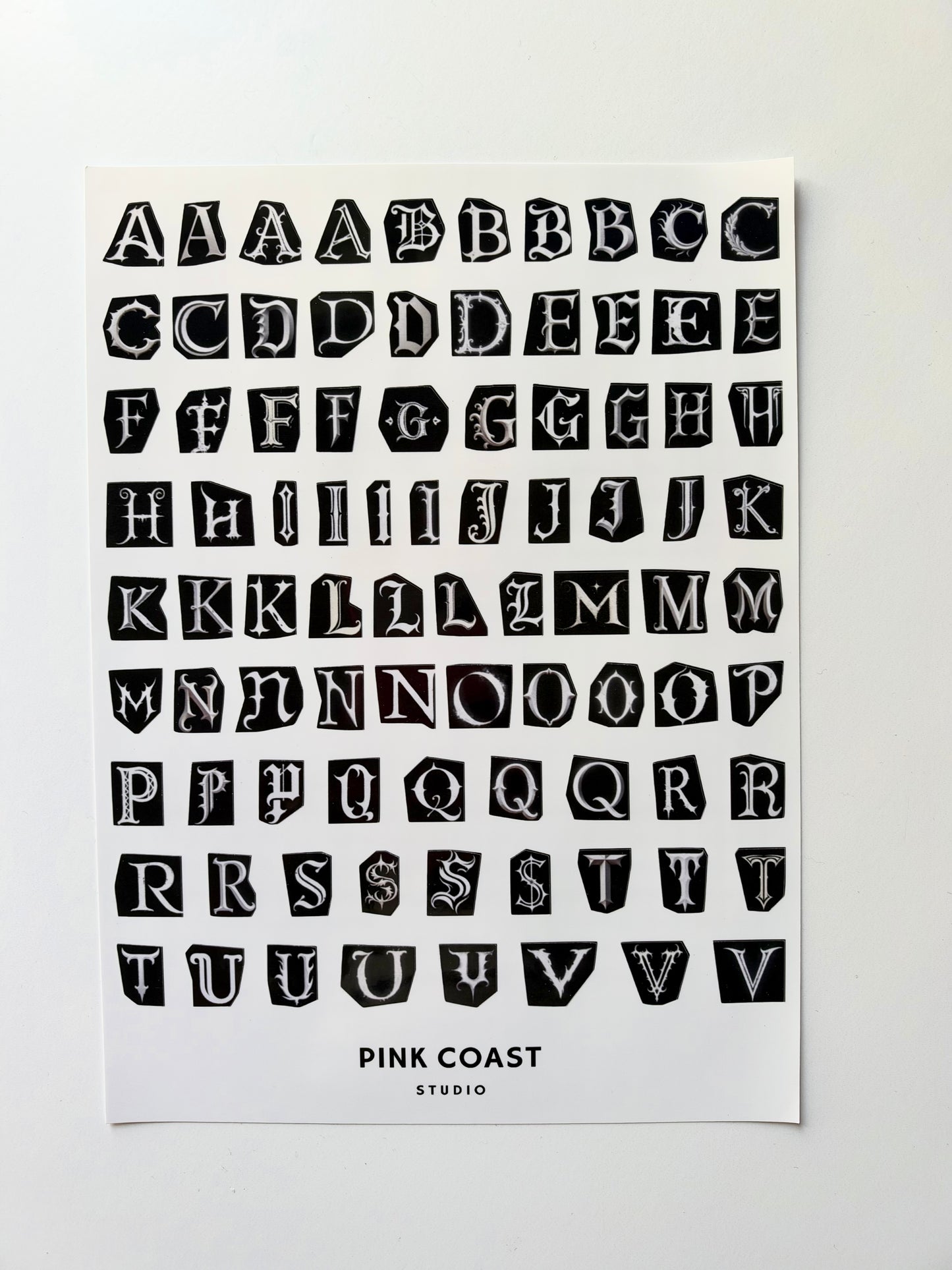 A typography art print featuring a black block-letter alphabet on a white sheet, with the text 'PINK COAST STUDIO' printed at the bottom, shown against a light gray background.
