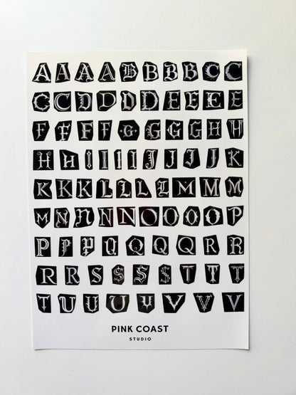 A typography art print featuring a black block-letter alphabet on a white sheet, with the text 'PINK COAST STUDIO' printed at the bottom, shown against a light gray background.