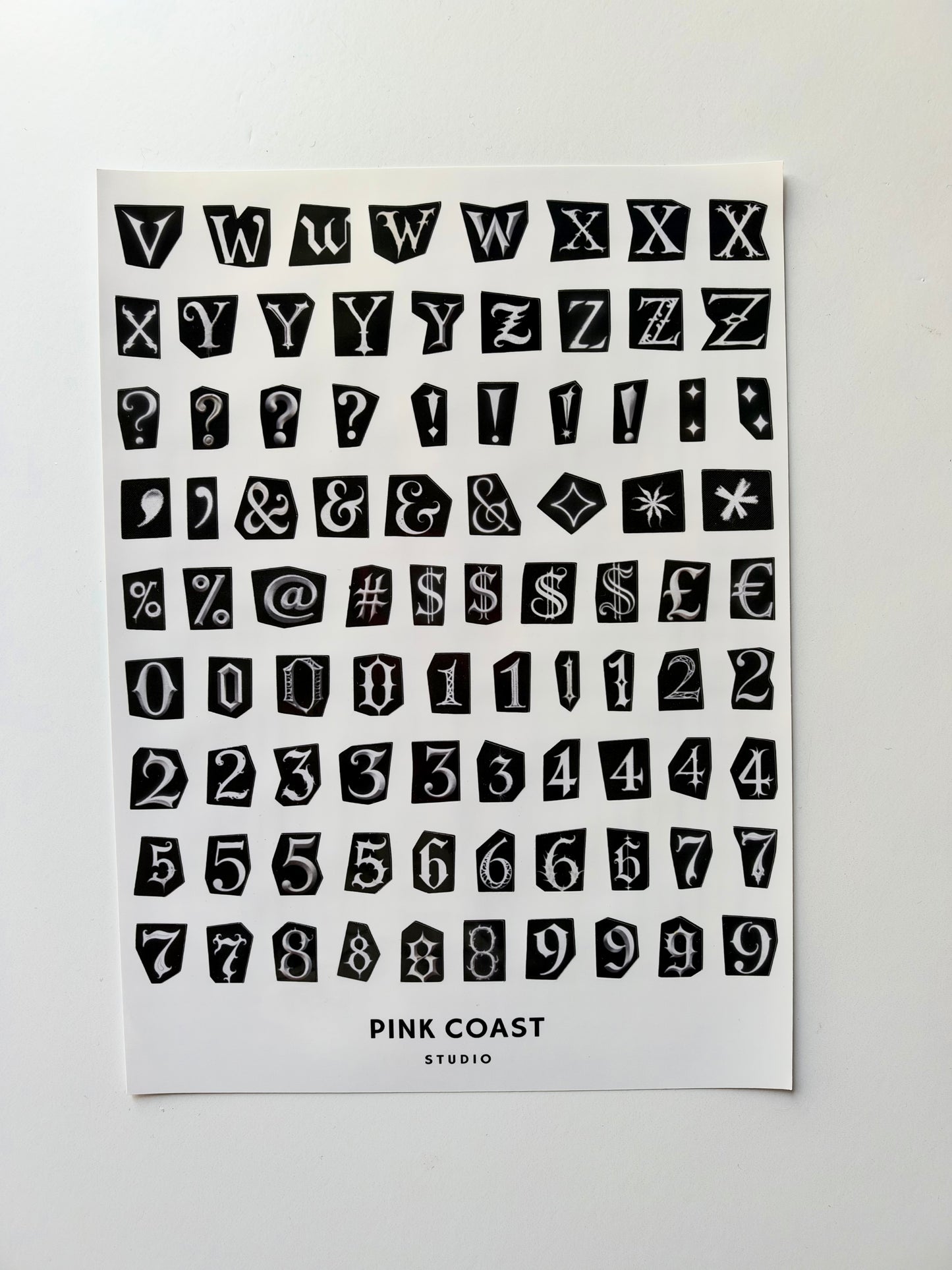 A sheet of black decorative block letters, numbers, symbols, and question marks on a white card, with the text 'PINK COAST STUDIO' printed at the bottom, photographed against a light gray background.