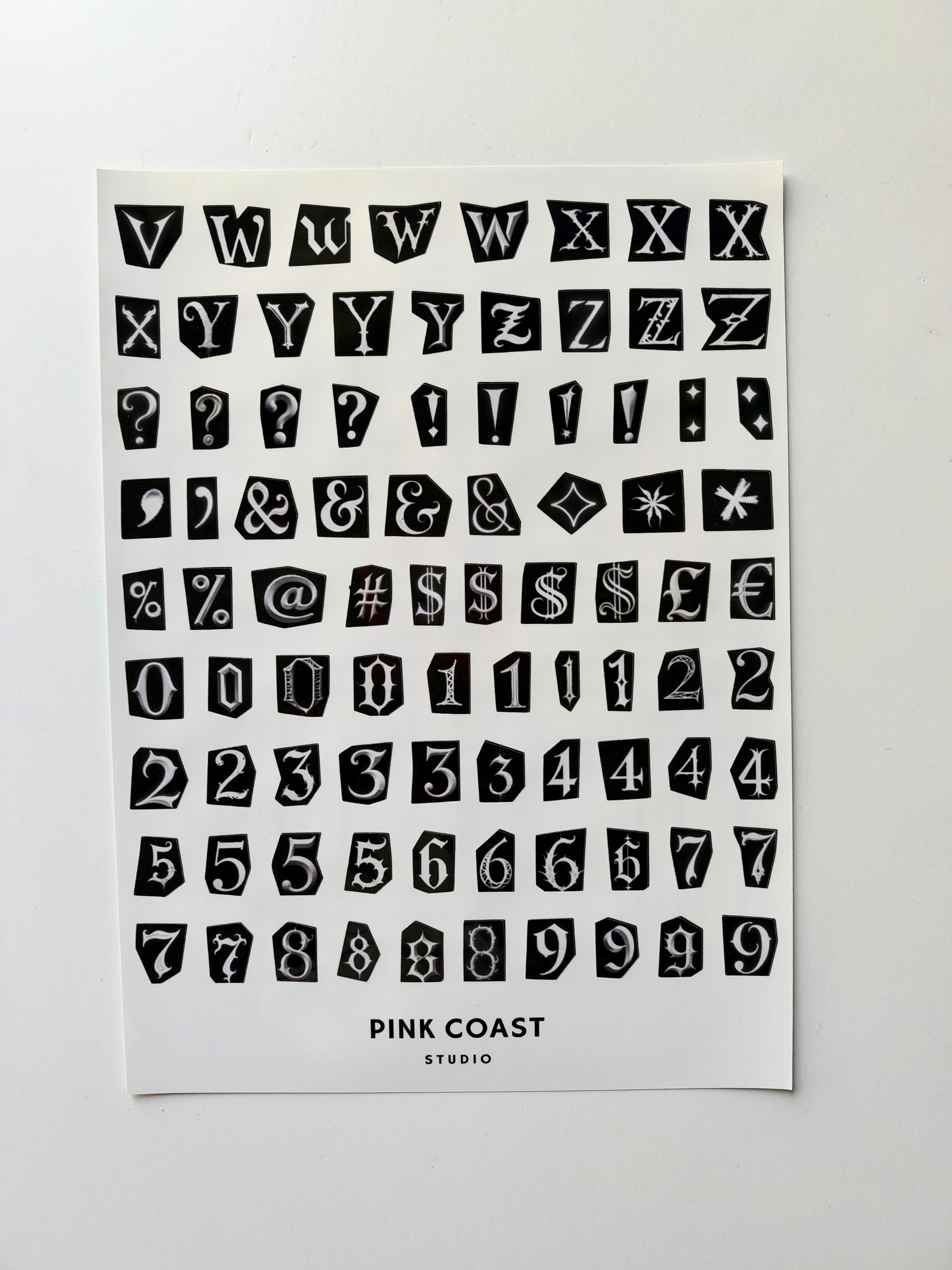 A sheet of black decorative block letters, numbers, symbols, and question marks on a white card, with the text 'PINK COAST STUDIO' printed at the bottom, photographed against a light gray background.