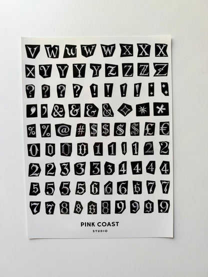 A sheet of black decorative block letters, numbers, symbols, and question marks on a white card, with the text 'PINK COAST STUDIO' printed at the bottom, photographed against a light gray background.