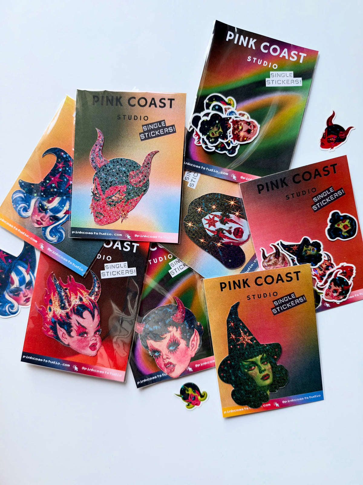 Several 'Pink Coast Studio' single sticker packs are arranged on a light gray background, with a few stickers placed in front of the packs. The packs feature colorful gradient covers with illustrated horror-themed faces, including a horned demon, a witch, and a vampire.