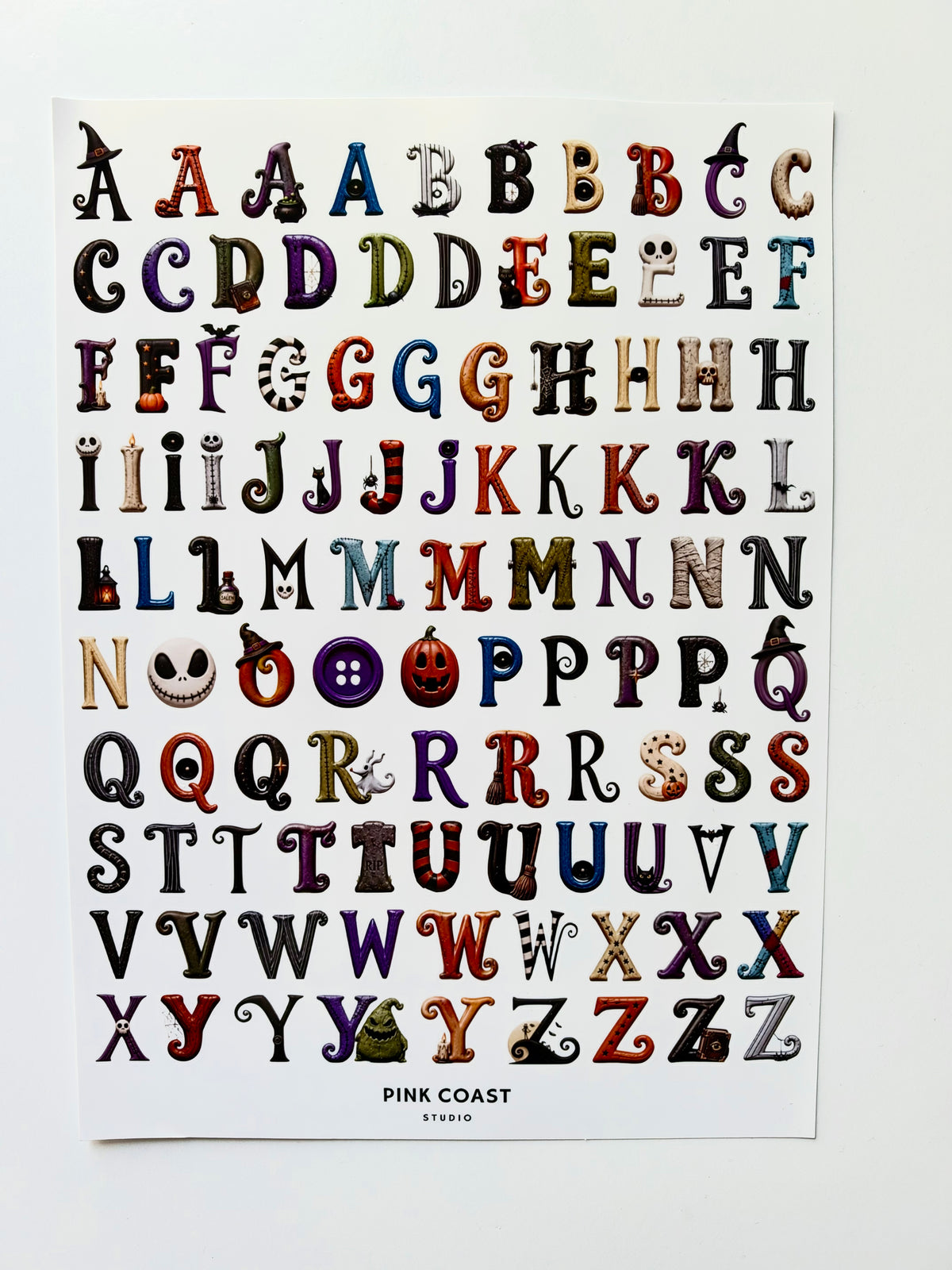 A printed alphabet sticker sheet featuring decorative, illustrated letters arranged in rows on a white sheet, with the text 'PINK COAST STUDIO' at the bottom, photographed against a light neutral background.