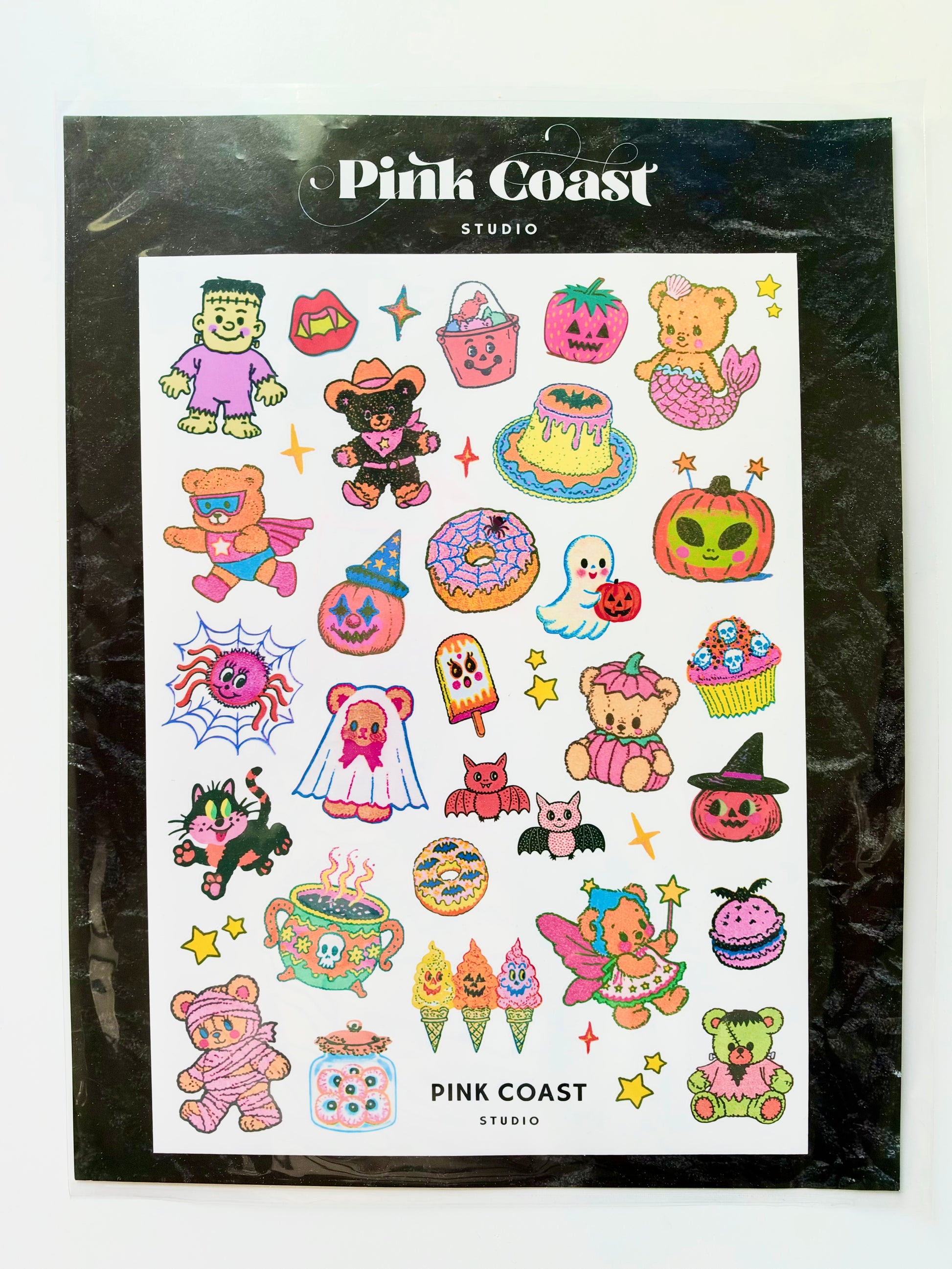 Pink Coast Studio Halloween-themed sticker sheet featuring cute cartoon characters and seasonal motifs like pumpkins, ghosts, teddy bears, cupcakes, and a mermaid, presented on a black backing card.